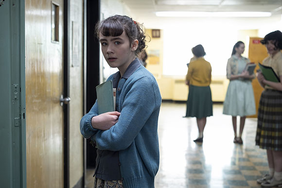 A young girl with dark hair and a blue cardigan stands in a school hallway holding a notebook.
