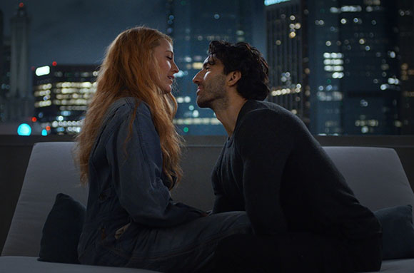 A man and a woman sit close together on a rooftop, facing each other against a city skyline at night.
