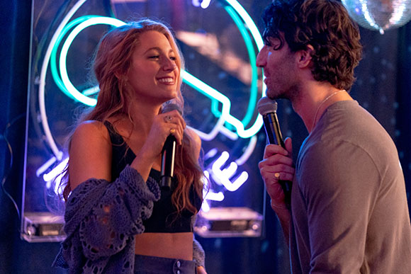 A smiling woman and a man hold microphones while performing in front of a glowing blue neon sign.