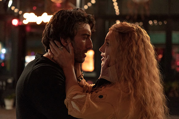A man and a woman with long wavy hair look intensely at each other in a dimly lit outdoor setting.