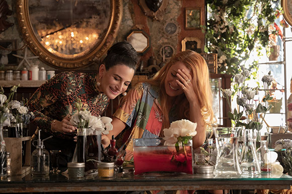 Two women laugh together while arranging white flowers in a cluttered, vintage-style flower shop.