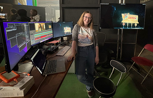 A woman stands in a studio next to a desk with multiple computer monitors and editing equipment.