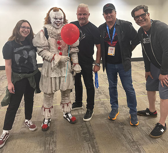 Five people posing in a room, including a person in a Pennywise clown costume holding a red balloon.