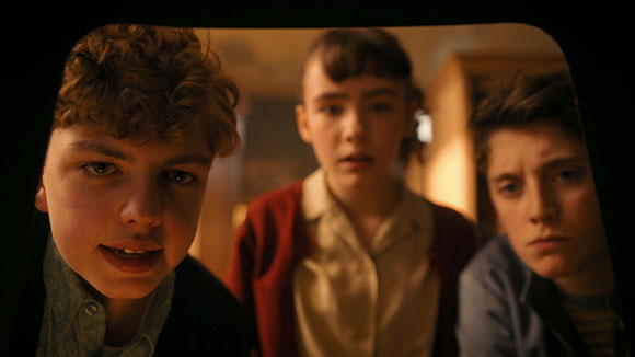 Three teenagers with serious expressions look directly into the camera in a dimly lit room.