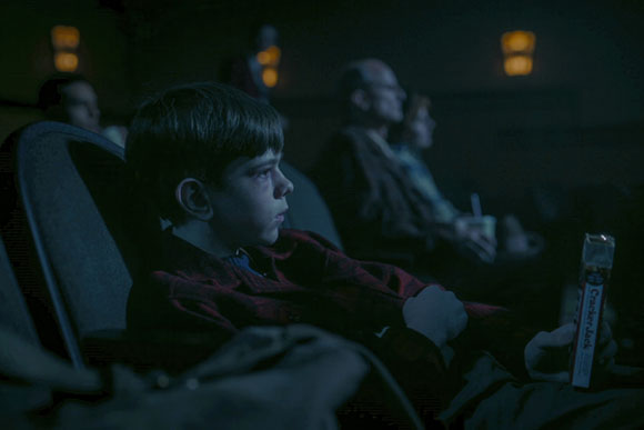 A young boy sits in a dark theater seat, looking forward while holding a snack.