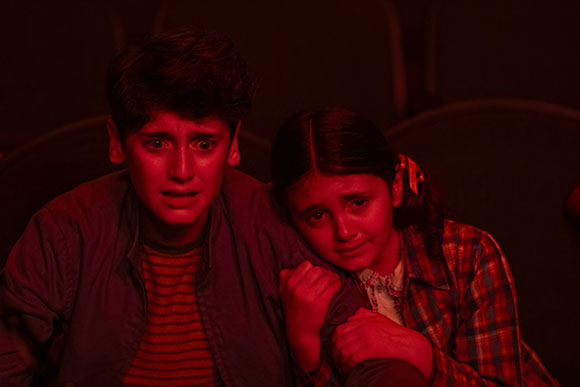 Two children with worried expressions sit together in dim, red-toned lighting.