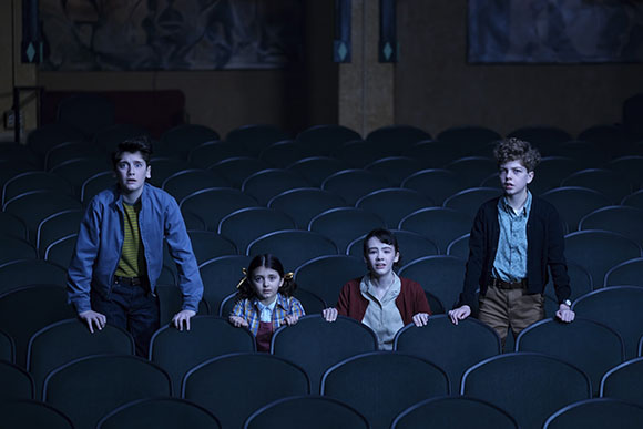 Four children sit in rows of empty dark theater seats, looking upward with surprised expressions.