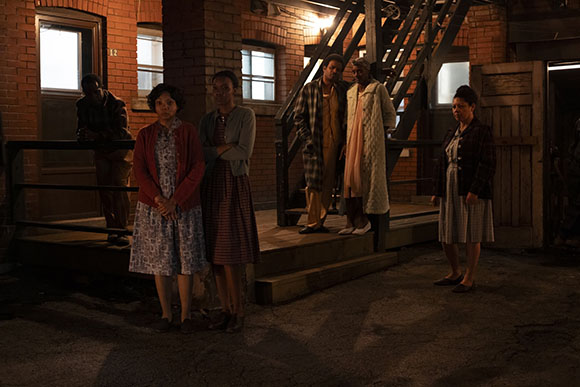 Five people stand in front of a brick building with a wooden staircase under dim lighting.