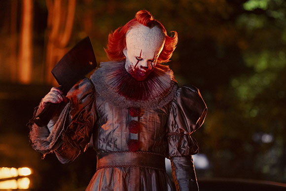 A clown with white face paint and red hair holds a large meat cleaver in a dark setting.