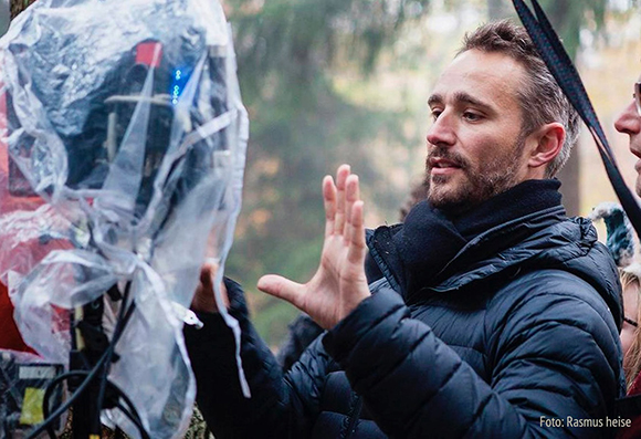 A man in a black puffer jacket gestures with both hands toward a camera wrapped in clear plastic.