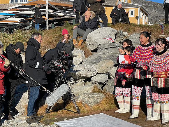 A film crew with a camera and microphone films a group of people in colorful traditional clothing near rocks.
