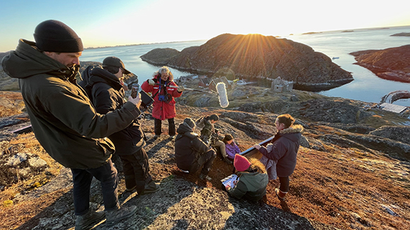 A group of people in winter clothing sit and stand on a rocky hill overlooking a coastal inlet at sunset.
