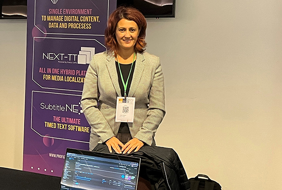 A woman in a grey patterned blazer and black top stands in front of a purple NEXT-TT banner.