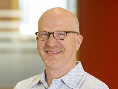 A bald man with glasses and a light-colored striped shirt smiling against a blurred background.
