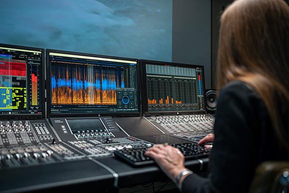 A person's hands typing on a keyboard while working at a large audio mixing desk with three computer monitors.