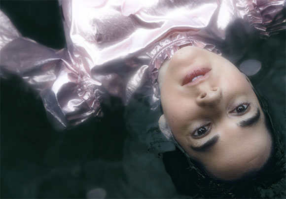 A person wearing a shimmering pink outfit floats upside down underwater against a dark background.