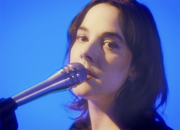 A woman with dark hair sings into a silver microphone against a blue background.