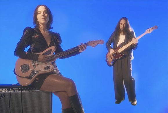 A woman sits on an amplifier playing a pink electric guitar while a man stands playing a bass guitar.