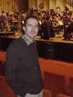 A man in a dark jacket stands in a theater with an orchestra performing on stage behind him.