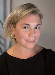 A woman with short blonde hair and gold hoop earrings wearing a black top.