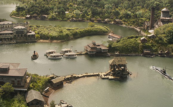 Aerial view of a village with wooden buildings, lush greenery, and boats on a winding river.