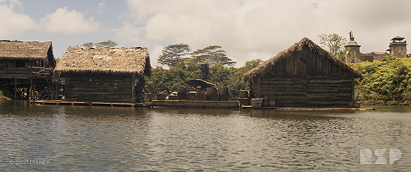 Several rustic wooden huts with thatched roofs sit along the edge of a calm body of water.