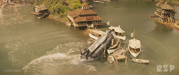 Aerial view of wooden buildings on a riverbank with several boats and a large dark vessel in the water.