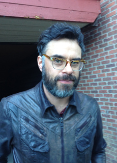 A man with dark hair, a beard, and glasses, wearing a leather jacket, stands in front of a brick wall.