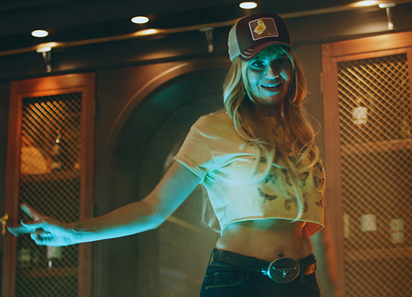 A blonde woman wearing a trucker hat and butterfly print crop top gestures with one hand.