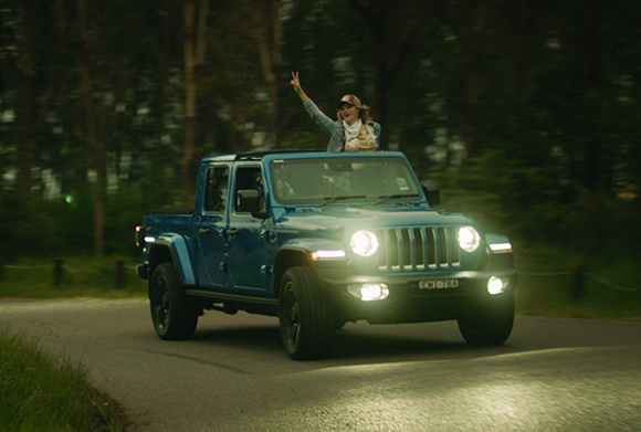 A woman in a hat waves from the bed of a blue moving pickup truck on a road through a forest.