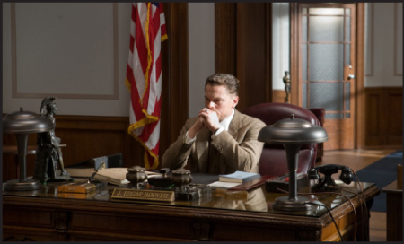 A man in a tan suit sits at a large wooden desk in a wood-paneled office next to an American flag.