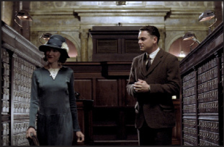 A man in a brown suit and a woman in a blue dress and hat stand between rows of wooden card catalog cabinets.