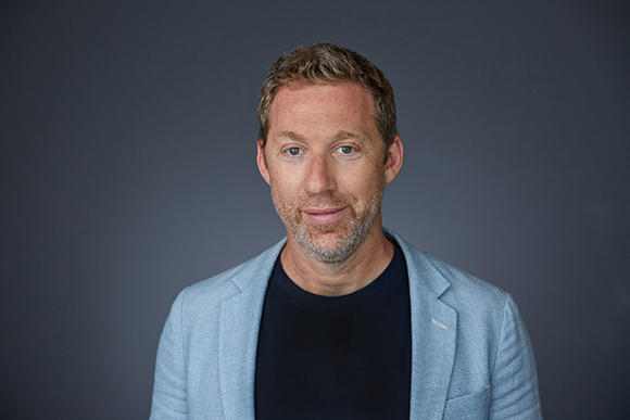 A man with short blond hair and stubble wearing a light blue blazer over a black shirt against a gray background.