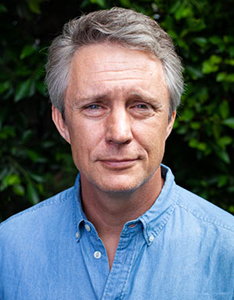 A middle-aged man with graying hair wearing a light blue button-down shirt against a leafy green background.