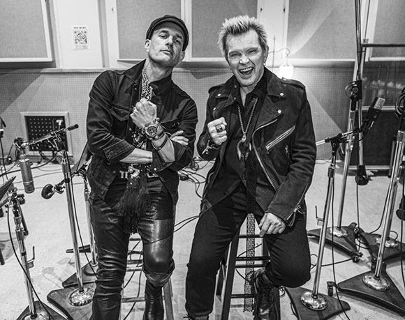 Two men in dark clothing pose with clenched fists in a recording studio surrounded by microphones.