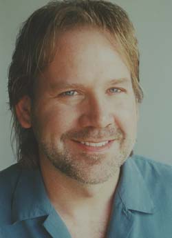 A smiling man with light brown hair and a short beard wearing a blue collared shirt.