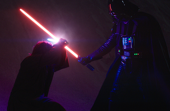 A figure in dark armor faces a kneeling figure, both wielding glowing red and blue lightsabers.
