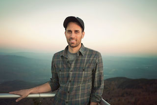 A smiling man wearing a dark baseball cap and a plaid button-down shirt stands before a hazy mountain landscape.