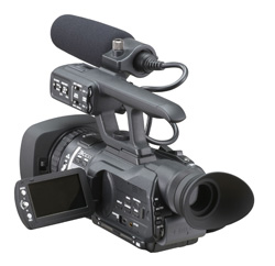 A black professional video camera with a top handle, shotgun microphone, and an open LCD screen.
