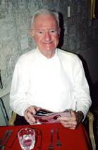 An older man with white hair wearing a white shirt sits at a table with a red tablecloth.