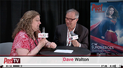 A woman in a red patterned shirt interviews a man in a dark suit at a table with microphones.