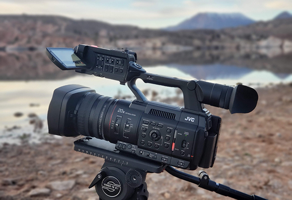 A black JVC professional video camera on a tripod facing a calm lake and distant mountains at dusk.