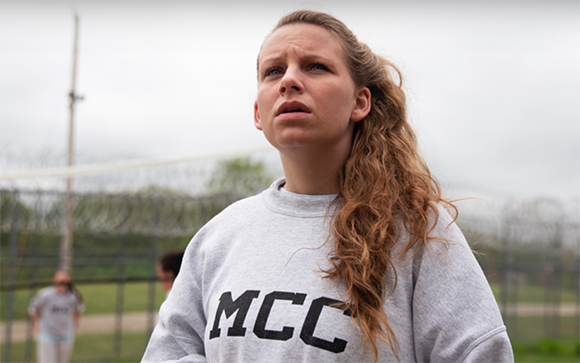 A young woman with long wavy hair wearing a grey MCC sweatshirt looks upward with a worried expression.