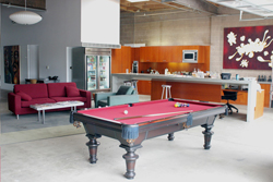 A red pool table in the foreground of a modern loft with a red sofa and a kitchen area.