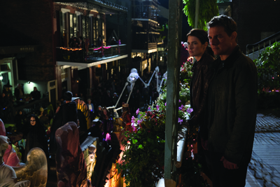 A man and woman stand on a balcony looking down at a crowded, dimly lit street with decorations.