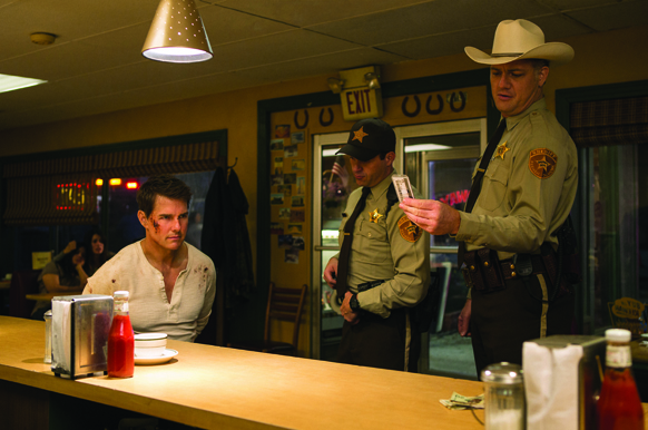 Two men in tan sheriff uniforms stand in a diner near a counter and an exit sign.