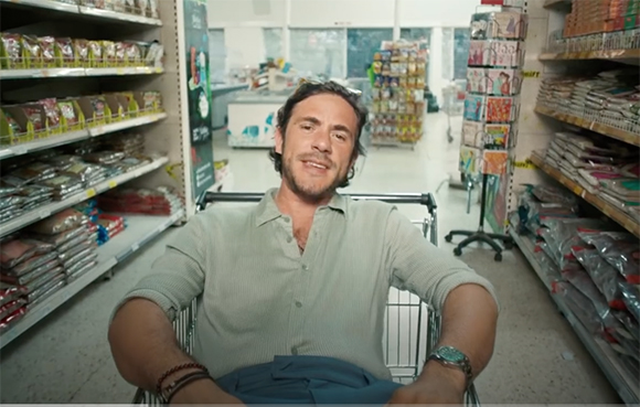 A man with dark hair and a light green shirt sits in a metal shopping cart in a grocery store aisle.