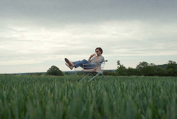 A person wearing sunglasses sits in a metal shopping cart placed in the middle of a green field under a cloudy sky.