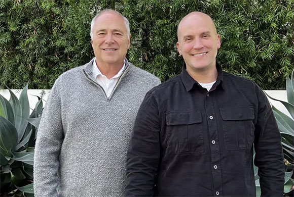 Two men smiling while standing together in front of green foliage and plants.