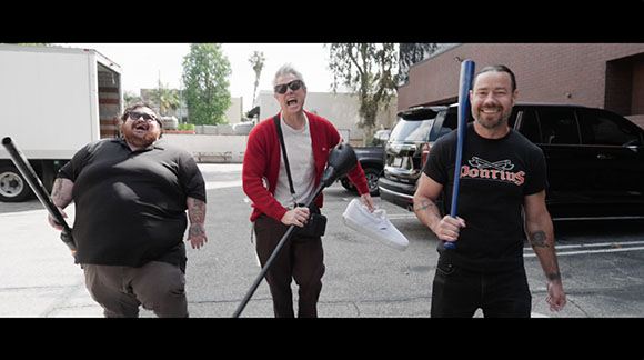 Three men smiling and holding baseball bats in a parking lot.